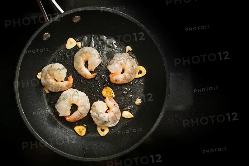 Fresh shrimps are sauteed with garlic in olive oil in a cooking pan on the black stove, preparation for a delicious appetizer, copy space, high angle view from above, selected focus