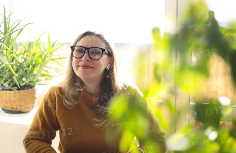 Portrait of smiling middle-aged woman wearing glasses sitting in room with green potted houseplants, selective focus. Positive happy 40s female teacher in eyeglasses in greenery, working in school