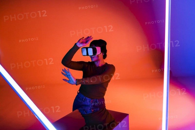 Happy female in VR headset smiling and exploring virtual reality under red neon illumination in futuristic room