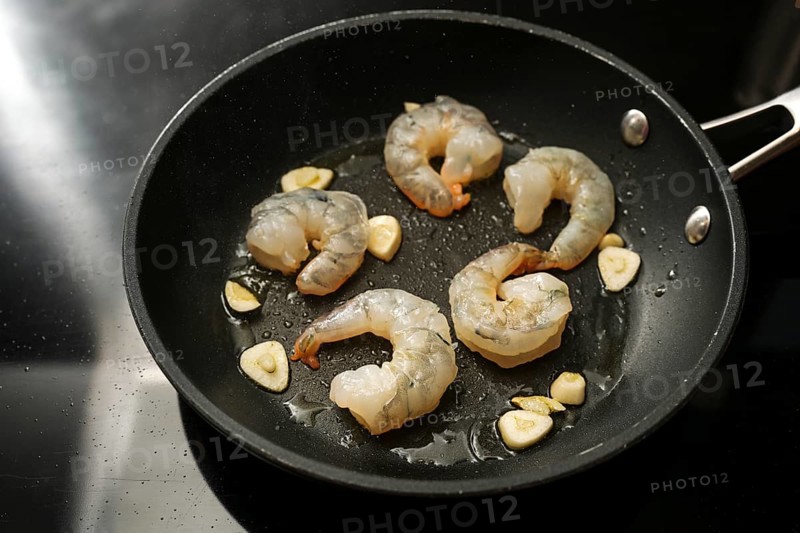 Raw peeled shrimps and garlic are sauteed in a cooking pan on the black stove, preparation for a delicious appetizer, copy space, selected focus, narrow depth of field