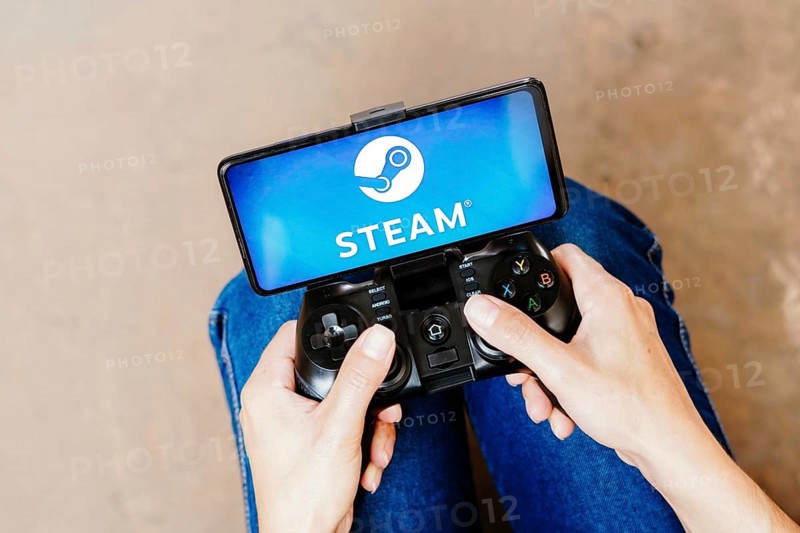 In this photo illustration, a person playing on a joystick and the Steam logo displayed on a smartphone screen