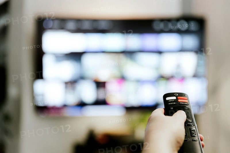 Hand holding remote control pointing to TV. Person watching smart television indoors. Blurred background. Colorful screen. Streaming series, channels. Woman browsing, looking for films online