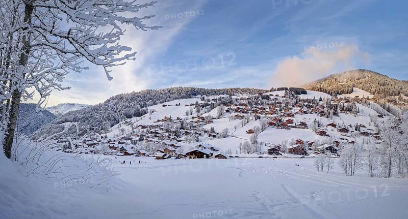 Winter panorama of snow covered La Clusaz village nestled in a hilly landscape in Annecy, France. Wooden cabins are scattered across the snowy hillside, surrounded by snow laden trees