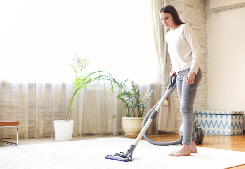 Full body young female using vacuum cleaner to tidy carpet in light room at home