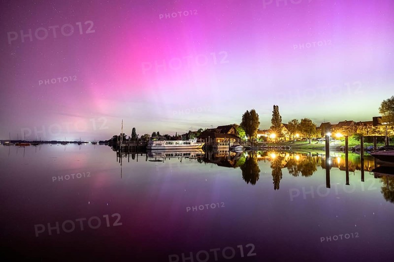 Colourful northern lights reflected in a quiet night lake next to illuminated buildings, harbour, Allensbach, Lake Constance, Baden-Württemberg, Germany, Europe