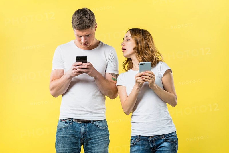 Suspicious girlfriend spying his boyfriend texting mobile phone against yellow backdrop