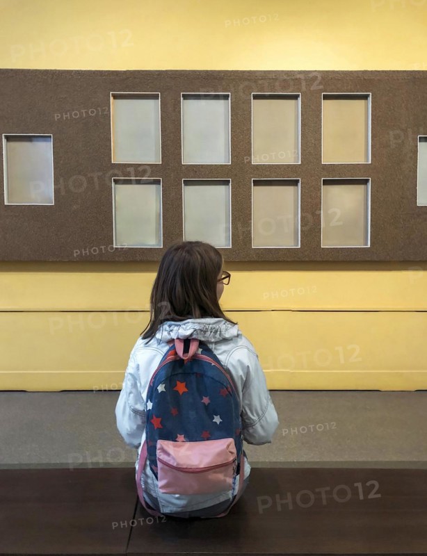 Back view of teen girl with backpack in museum visiting exhibition, schoolgirl sitting on bench in art gallery and looking with interest at art objects or exhibits on wall