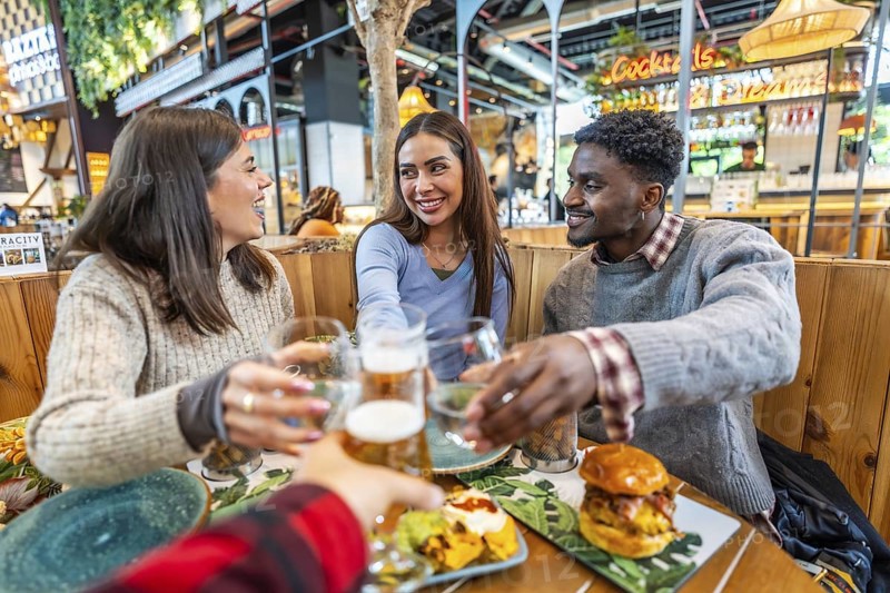 Group of cheerful multi ethnic friends toasting with beer and cocktails, enjoying meal together at food market