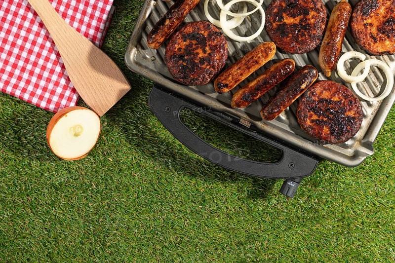 Barbecue with sausages and burgers next to a red and white chequered cloth on a green lawn