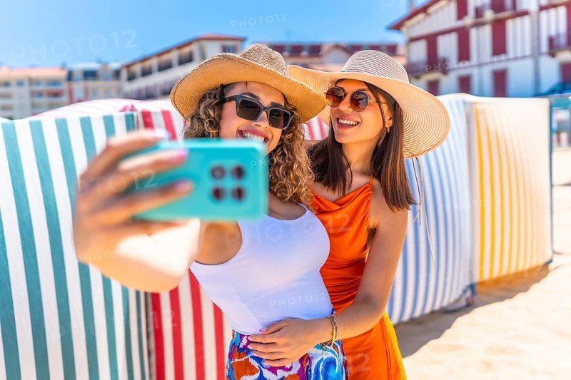 Two smiling women with sun hats and sunglasses taking a selfie on a sunny beach with colorful striped beach huts