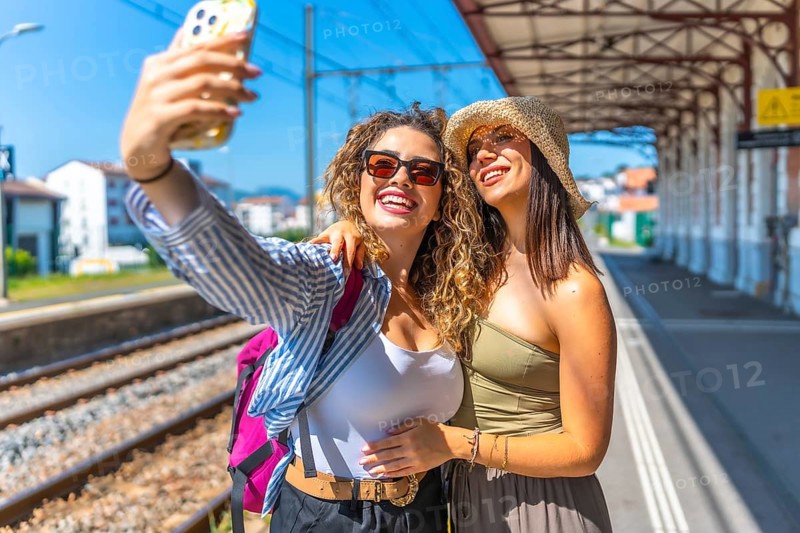 Two happy female tourists taking a selfie with smartphone at a train station, enjoying their summer vacation