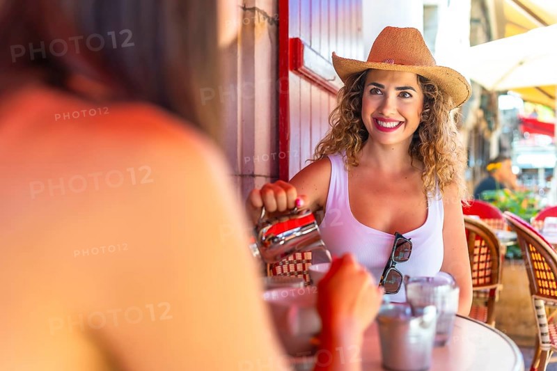 Smiling tourist wearing straw hat pouring milk into coffee, having breakfast with friend at outdoor cafe