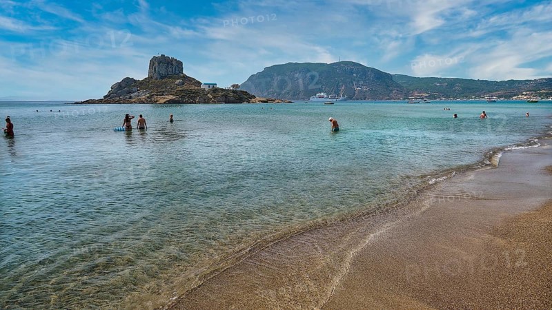 Clear sea with people bathing, surrounded by an island and mountains under a cloudy sky, Agios Stefanos Beach, Agios Stefanos Basilica, Chapel of Agios Nikolaos, Kastri Island, Kos, Dodecanese, Greek Islands, Greece, Europe