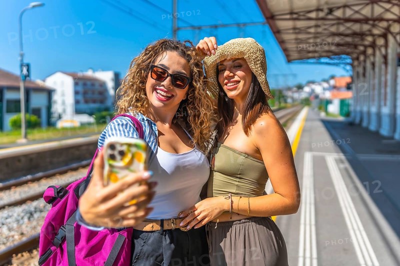 Two female tourists taking a selfie with smartphone on a train station platform during summer vacation