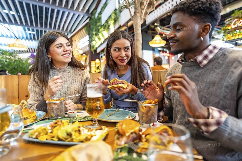 Group of happy multiethnic friends enjoying a meal together, eating burgers, fries and drinking beer, sitting at a restaurant table inside a food court