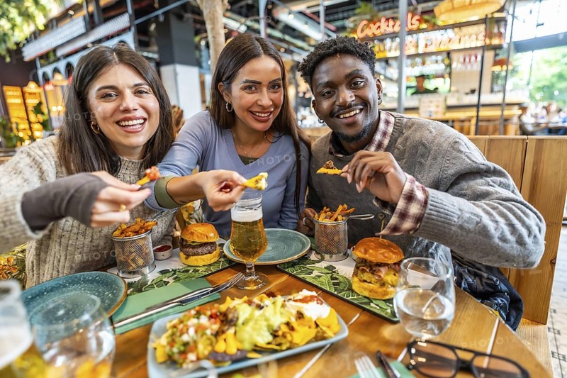 Three cheerful multi ethnic friends are enjoying a meal together, sharing nachos, burgers, fries, and beer in a vibrant restaurant setting