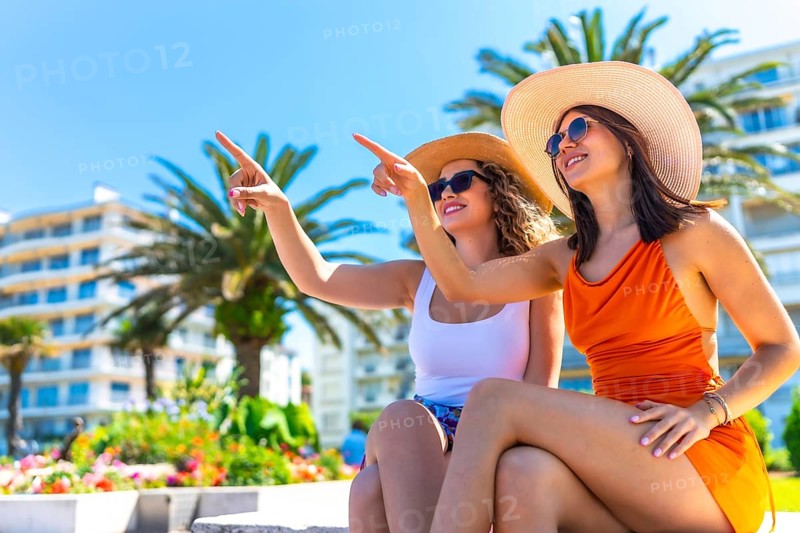 Two happy female tourists wearing sunglasses and summer hats, pointing their fingers at something while enjoying their vacation