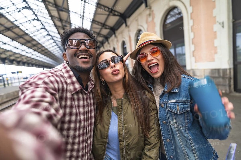 Three happy young tourists are taking a selfie on a train station platform, excited for their next adventure
