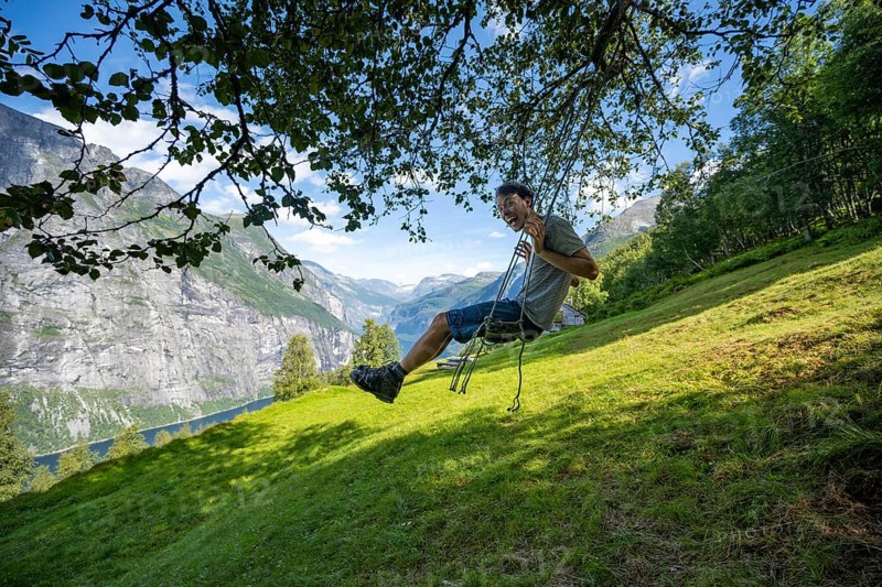 Young man swinging, Blomberg Gård, above the Geirangerfjord, view of idyllic fjord landscape, near Geiranger, Møre og Romsdal, Norway, Europe