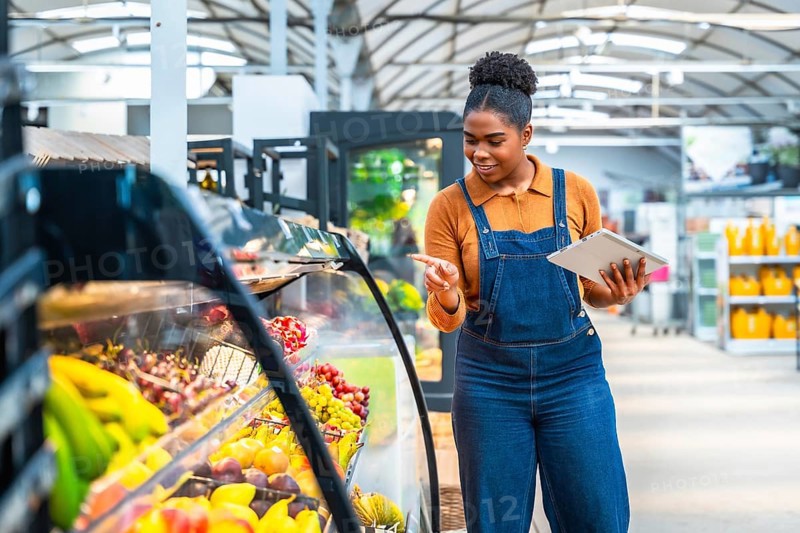Young supermarket employee using a digital tablet and checking fresh products in a refrigerated display case