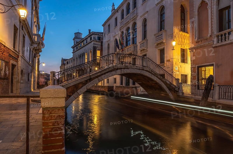 Small pedestrian bridge arching over canal in venice, italy at twilight with light trails reflecting in water from passing boat