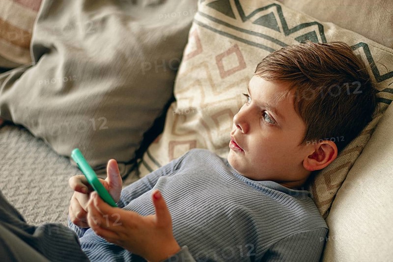 A boy holds a smartphone in his hands, relaxing on the couch, in a cozy living room, natural light, lifestyle