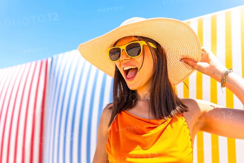 Carefree young woman wearing a straw hat and sunglasses enjoying summer vacation against a colorful striped background