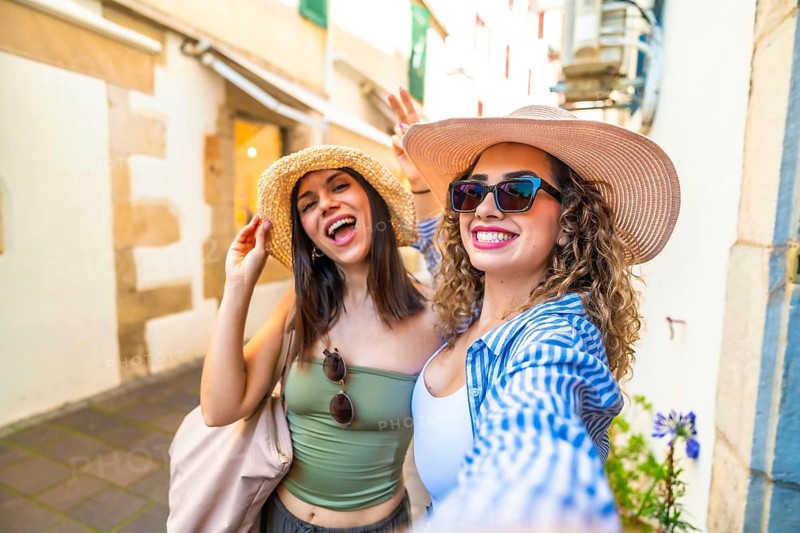 Two joyful women with straw hats and sunglasses are taking a selfie with a cheerful expression, enjoying their summer vacation in a picturesque european town