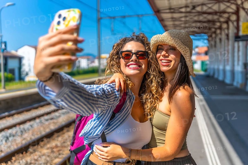 Two female tourists with backpack and straw hat are taking a selfie with smartphone at a train station platform