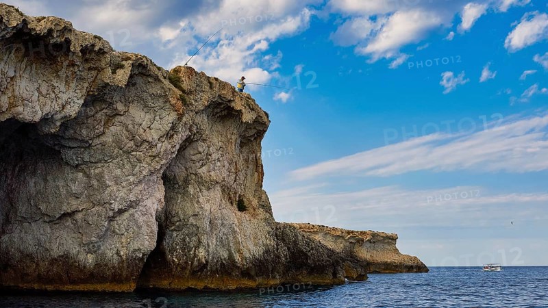 Person fishing from a rocky cliff under a cloudy sky, boat trip around the island of Ortigia, Isola di Ortigia, Syracuse, Sicily, Italy, Europe