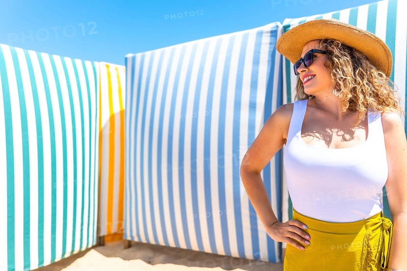 Young woman with curly hair, sunglasses and straw hat posing on beach with colorful striped cabanas in background during sunny summer day