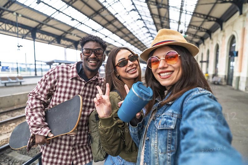 Happy multi ethnic friends taking a selfie at the train station before departure, holding skateboard and reusable water bottle