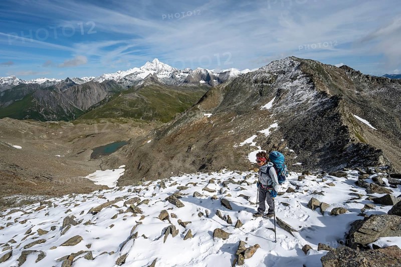 Mountaineer in boulder terrain with fresh snow in summer, ascent to the summit Böses Weibl, view of the mountain landscape with Kesselkeessee, snow-covered summit of the Großglockner in the background, Wiener Höhenweg, Schober group, Hohe Tauern National Park, Carinthia, Austria, Europe