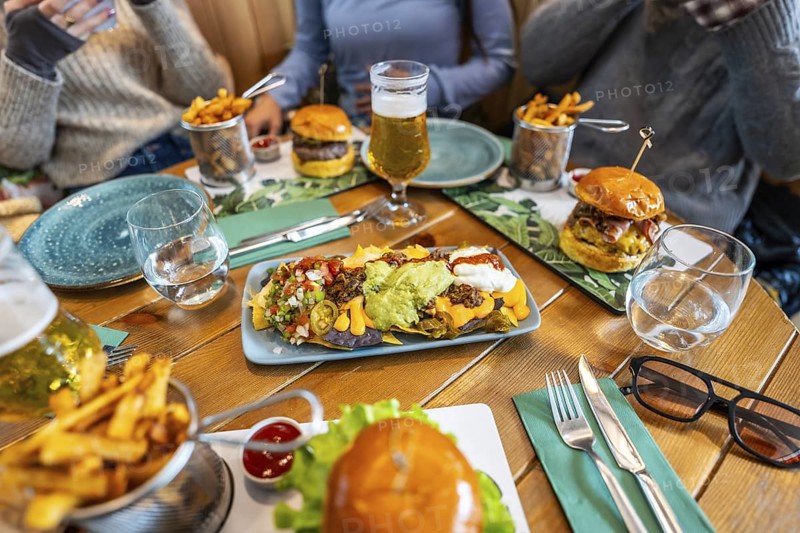 Plate of loaded nachos takes center stage on a restaurant table, surrounded by burgers, fries, and beers, as friends gather for a casual meal
