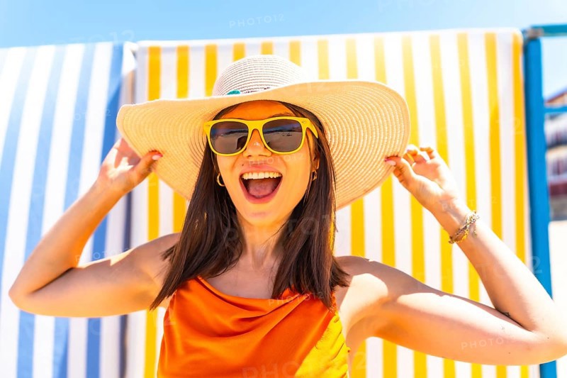 Young woman wearing a straw hat and sunglasses, smiling brightly while enjoying the summer sun, holding her hat with joy and excitement