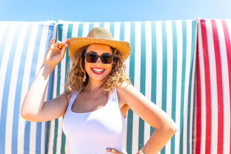 Cheerful young woman with curly hair, wearing sunglasses and a straw hat, smiles while posing confidently against a backdrop of vibrant striped beach towels, enjoying a sunny day at the beach