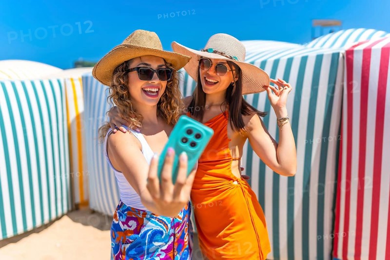 Two happy women wearing summer hats and sunglasses are taking a selfie with a smartphone on a sunny beach, near colorful beach huts