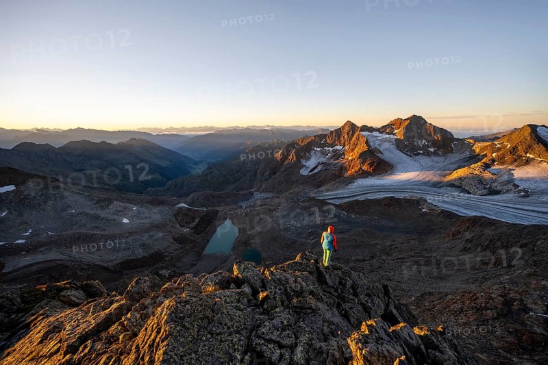 Mountaineer in front of picturesque high mountain landscape at sunrise with alpenglow, blue glacial lakes, glaciers and rocky mountain peaks in the morning light, view at the Becher on mountain peaks Königshofspitz and Sonklarspitze and Schwarzwandspitz with glacier Übeltalferner, Stubai Alps, South Tyrol, Italy, Europe