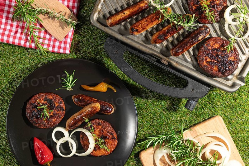 Grilled sausages and burgers on a barbecue and plates with rosemary on a green lawn