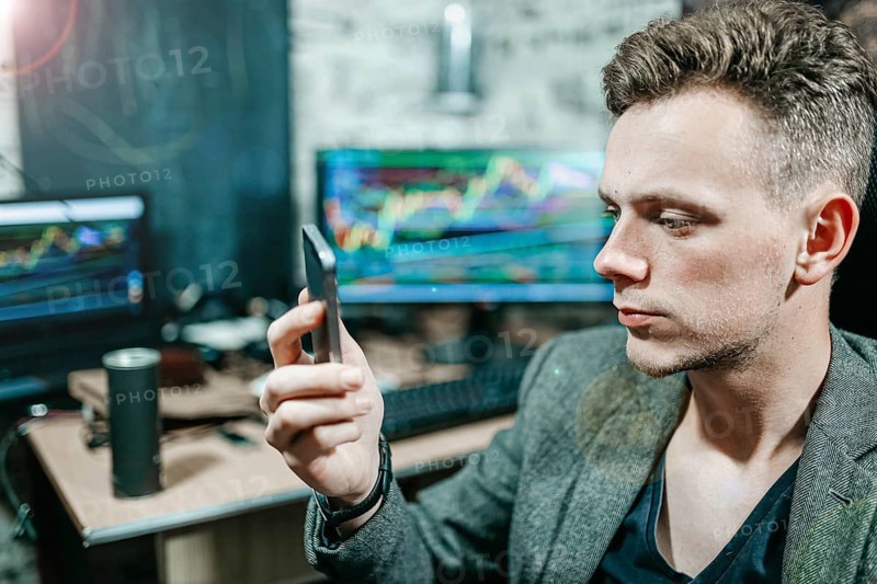 Man focused on a smartphone in an office with trading charts on monitors