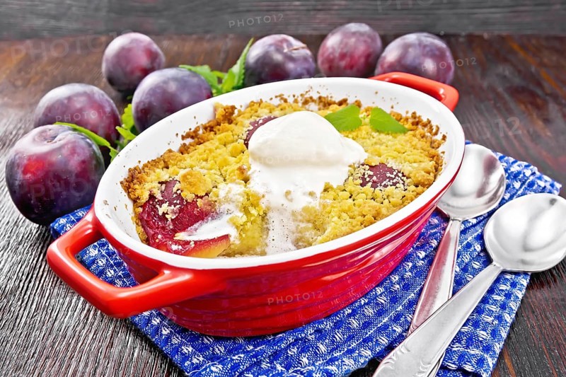 Crumble with plum and ice cream in a red brazier with spoons on a napkin, plums and mint on dark wooden board background