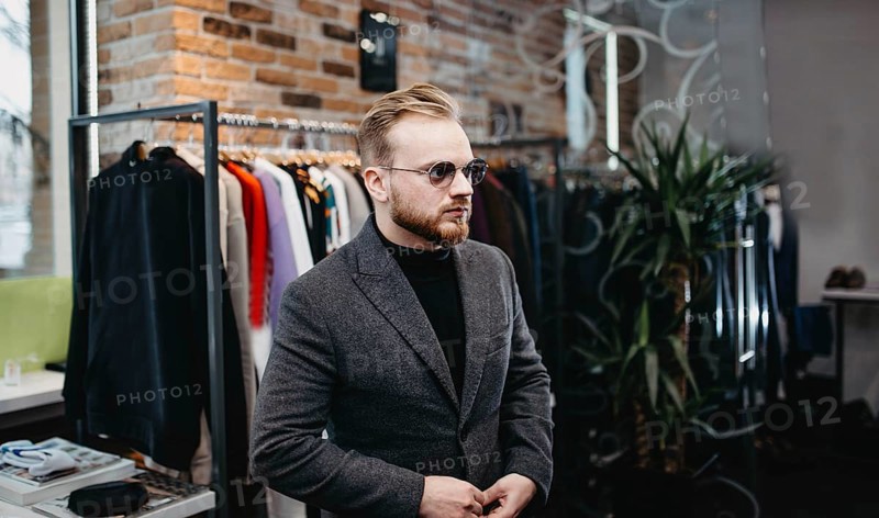 Man wearing sunglasses and a coat stands inside a chic clothing store with trendy decor