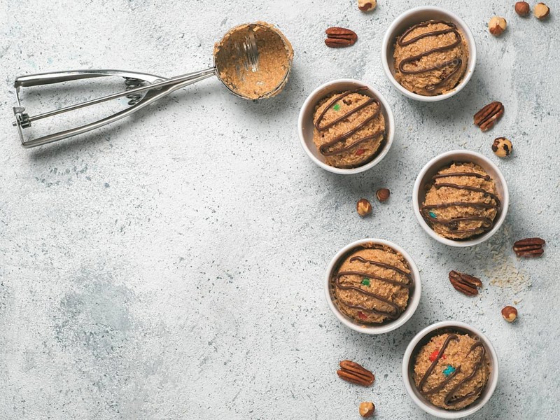 Safe-to-eat raw monster cookie dough in small portion bowl, ice cream scoop and nuts on gray cement background. Ideas and recipes for kids and toddlers meal. Top view or flat-lay. Copy space for text
