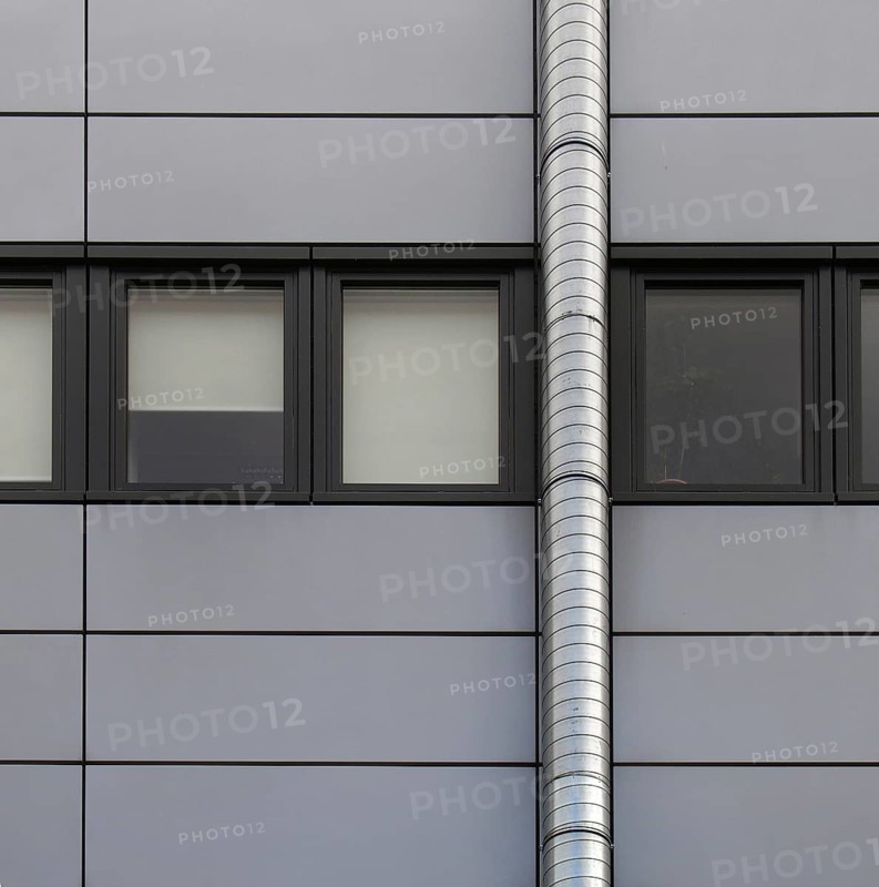 Grey cladding panels on an industrial building with a row of back window frames and a silver pipe running up the wall