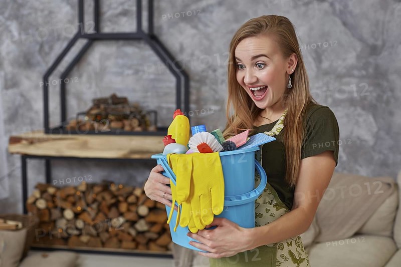 A cleaning woman is standing in living room holding a blue bucket fulfilled with chemicals and facilities for tidying up in her hand. Cleaning service