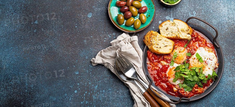Middle Eastern and Maghrebi healthy dish Shakshouka made of eggs and tomato sauce served in pan with toasts, fresh cilantro and olives on rustic concrete background table from above, space for text, food photography, food photography