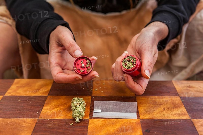 Selective focus closeup of person's hands holding a red weed filled grinder shot from above. Checkered table with rolling paper, filter and bud displayed below