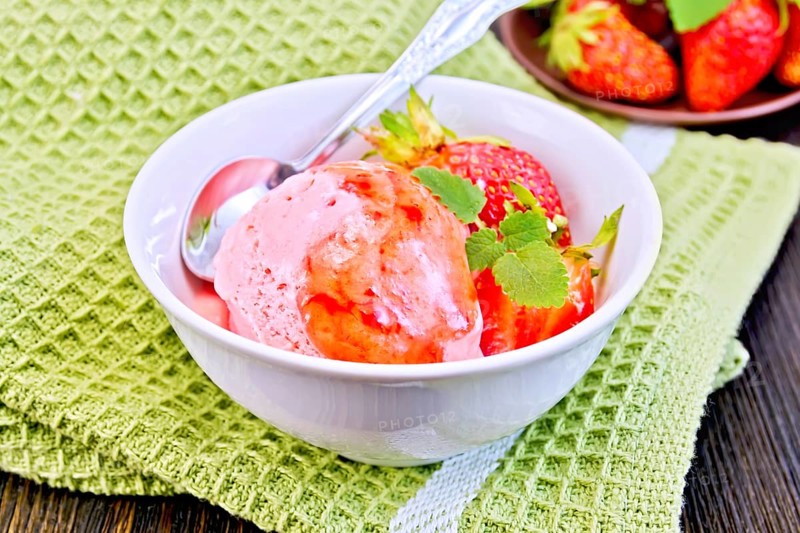 Strawberry ice cream in a white bowl with strawberries, strawberry syrup and a spoon on a napkin on a wooden boards background