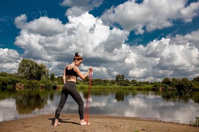 A woman exercises using a resistance band by a tranquil river, with dramatic clouds above