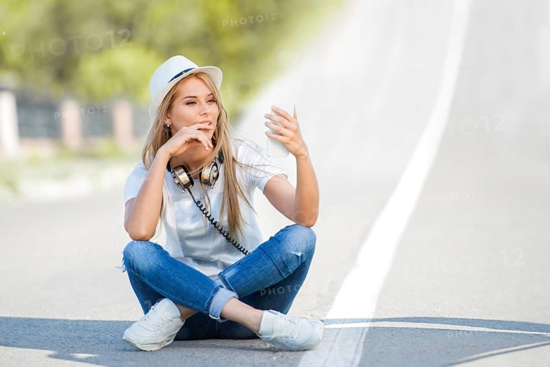 Happy young woman with vintage headphones around her neck, surfing the internet on her smartphone and sitting on a divider against the backdrop of a street